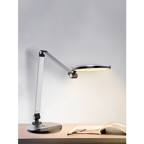 Een zilveren bureaulamp met een ronde lampkop die een gelige verlichting geeft. Op een bureau naast een open boek.