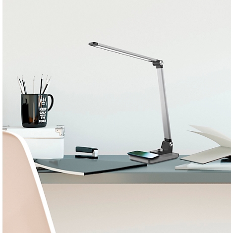 Eine silberne Schreibtischlampe steht auf einem Schreibtisch. Auf dem Schreibtisch sind auch eine Tasse, Hefte und Papier zu sehen.
