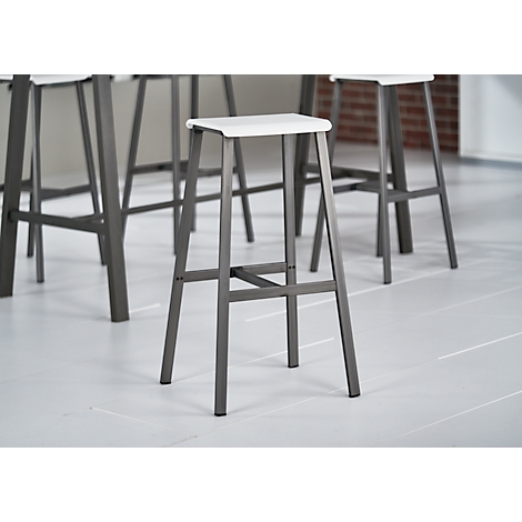 A bar stool with a white seat and gray metal frame. Several stools are standing in front of a white floor and a red brick wall.