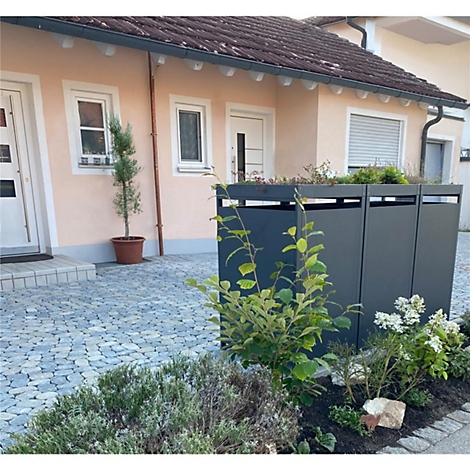 Donkergrijze vuilnisbakkenkast voor een lichtroze huis, omgeven door planten. Kasseien aan de voorkant.