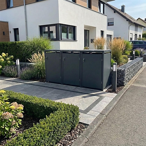 Drie grijze vuilnisbakken voor een huis. De dozen zijn omgeven door groene planten. Op de achtergrond is een huis met moderne architectuur te zien.