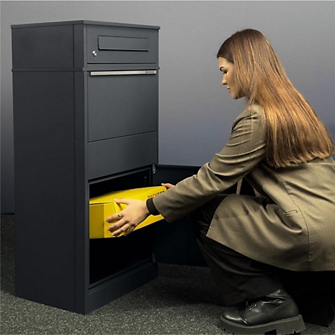 Vrouw hurkt voor een grijze pakketbox en haalt een geel pakket eruit. Lang bruin haar, donkerbruine jas.