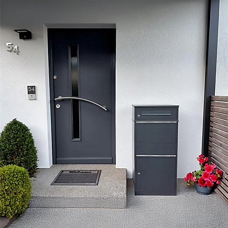 Porte d'entrée gris foncé avec insert en verre, à côté une boîte aux lettres. Devant la porte, un paillasson et de petits buissons.