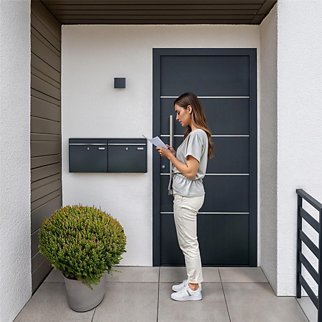 Femme devant la porte, lisant une lettre. Porte sombre, boîtes aux lettres grises, pot avec plante, pantalon et chaussures blanches.