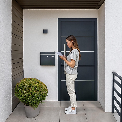 Femme lisant des lettres devant une porte. Porte grise, boîte aux lettres grise. Plante en pot devant.