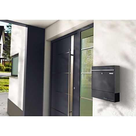 Boîte aux lettres noire fixée au mur à côté d'une porte d'entrée sombre. La photo a été prise à l'extérieur.