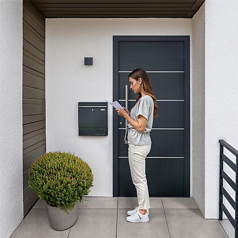 Une femme se tient devant la porte d'entrée, lisant des lettres. A côté d'elle, une boîte aux lettres. Au premier plan, un buisson en pot.