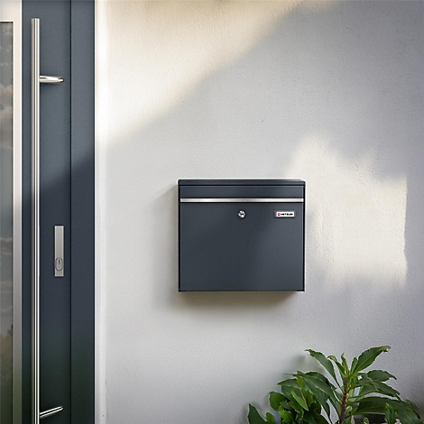 Dunkelgrauer Briefkasten an weißer Wand, unterhalb grüne Pflanzen. Daneben eine graue Tür.