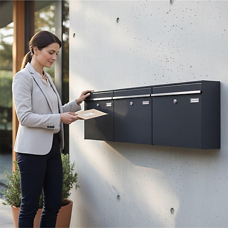 Eine Frau öffnet einen Briefkasten, der an einer Betonmauer befestigt ist. Sie trägt einen Blazer, eine Hose und hält den Brief in der Hand.