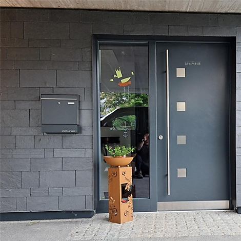 Dunkelgraue Haustür mit Edelstahlapplikationen und Briefkasten an einer Steinwand. Vor der Tür steht eine Pflanzsäule.