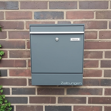 Grauer Briefkasten an einer Backsteinmauer, Aufschrift "Zeitungen".