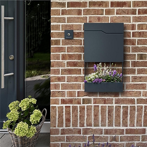 Dunkelgraue Briefkasten mit integriertem Blumenkasten an einer Backsteinmauer. Daneben eine Tür und ein Korb mit Blumen.