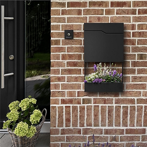 Schwarzer Briefkasten mit integriertem Blumenkasten an einer Backsteinmauer, neben einer Tür und einem Korb mit Blumen.