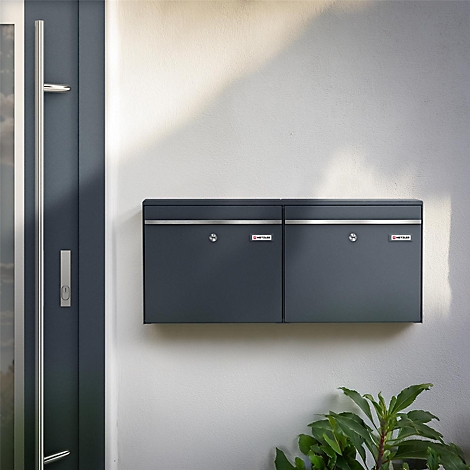 Donkergrijze dubbele brievenbus op een witte muur. Daarnaast een deur. Groen gebladerte rechtsonder.
