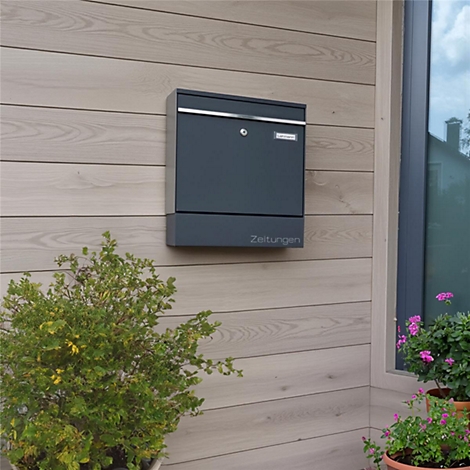 Donkergrijze brievenbus op houten gevel. Inscriptie "Kranten". Rechts een raam met uitzicht op het groen.