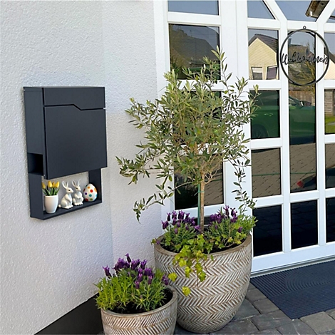 Een antracietkleurige brievenbus met plank, paasdecoratie en bloeiende planten voor een witte deur.