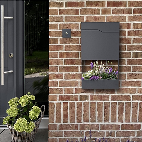 Donkergrijze brievenbus met bloembak aan bakstenen muur. Daarnaast voordeur en mand met bloemen. Deurbel boven de doos.