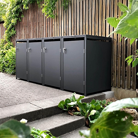 Graue Mülltonnenboxen vor Holzzaun. Grüne Pflanzen umgeben die Boxen.