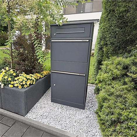Dunkelgrauer Briefkasten mit drei Fächern, steht auf Kies neben Blumen und Büschen.