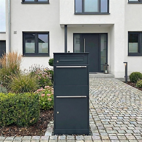 Dunkelgraue Briefkastenanlage mit zwei Fächern und einem separaten Fach darüber. Sie steht vor einem Haus mit Garten.