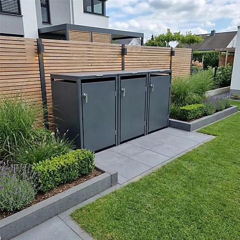 Drie grijze vuilnisbakken staan in een tuin voor een houten hek, omringd door planten en gazon.