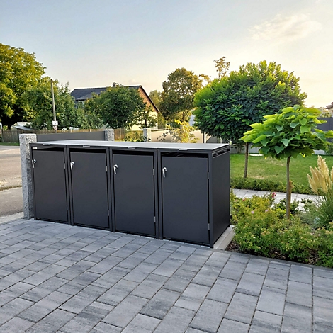 Dunkle Mülltonnenbox vor einem Haus. Geplasterte Fläche, grüne Bäume, blauer Himmel im Hintergrund.