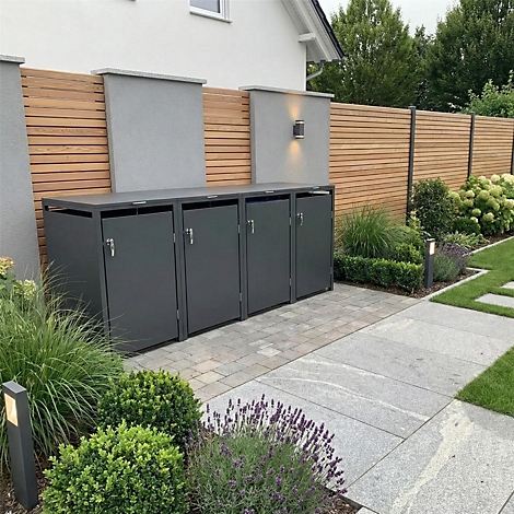 Graue Mülltonnenbox vor Holzlattenzaun. Davor Garten mit Pflanzen und Weg. Im Hintergrund Haus.