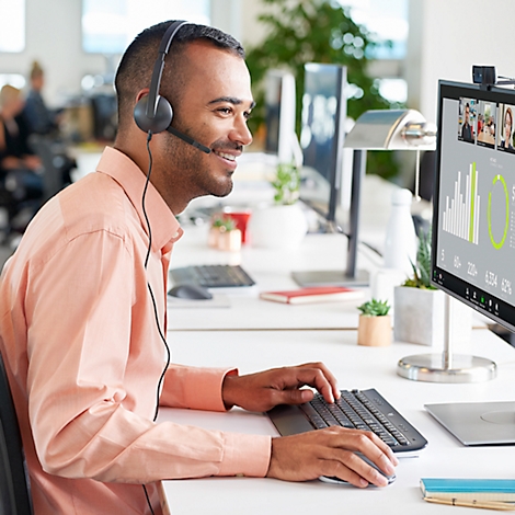 Un homme souriant avec un casque est assis à un bureau, regardant son ordinateur et travaillant. Un appel vidéo est visible à l'écran.