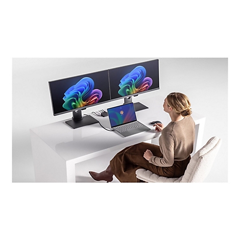 Eine Frau sitzt vor einem Schreibtisch mit zwei Monitoren und einem Laptop. Die Monitore und der Laptop zeigen bunte Grafiken.