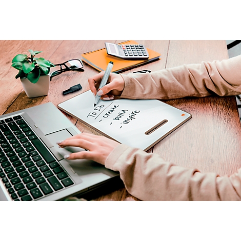 Une personne écrit avec un stylo sur un bloc-notes, un ordinateur portable et d'autres accessoires sur une table en bois.