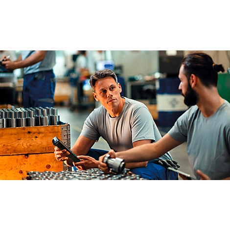 Zwei Arbeiter in einer Fabrik prüfen Metallbauteile und nutzen ein mobiles Scan-Gerät.