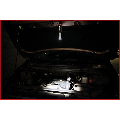 Ein offener Motorraum eines Autos wird von einer Stablampe ausgeleuchtet. Die Lampe hängt an der Motorhaube.