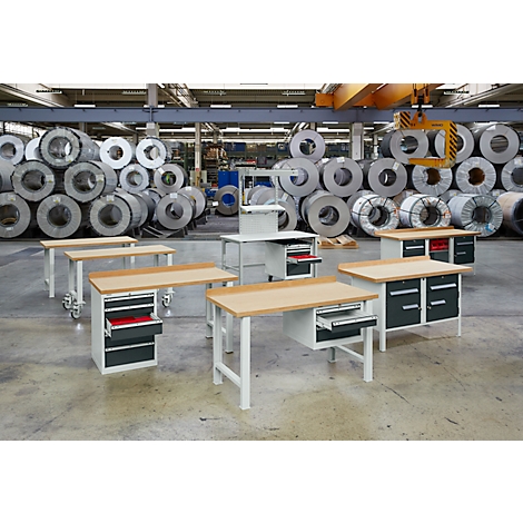 Workbenches in a factory hall. Behind the tables are large metal rolls stacked.