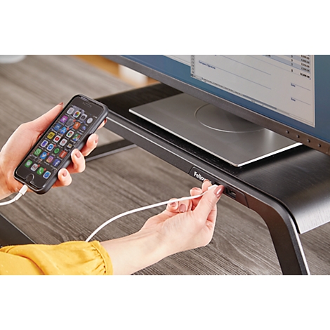 Person connects smartphone with charging cable to a monitor stand. Screen with applications, yellow top.