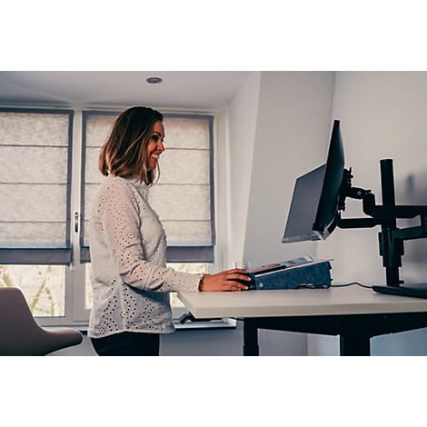Eine lächelnde Frau steht an einem Stehpult und arbeitet, ein Monitor ist im Hintergrund sichtbar.