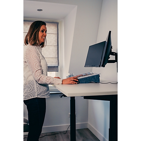 Frau steht lächelnd an einem höhenverstellbaren Schreibtisch mit Computer. Fenster im Hintergrund.