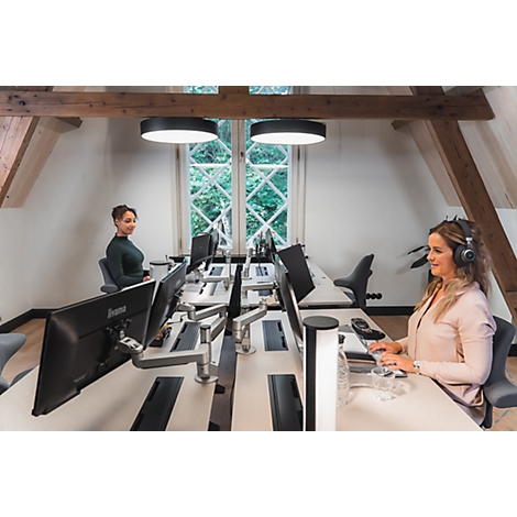 Zwei Frauen in einem Büro mit Schreibtischen, Computern und Kopfhörern. Helles Büro mit Holzbalken und Fenstern.