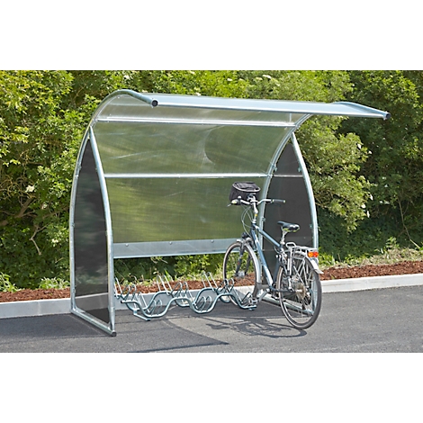 Bicycle stands under a transparent shelter with a metal frame. Trees in the background.