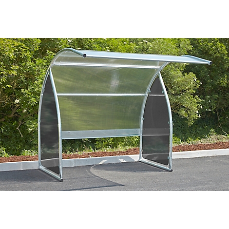 Metal bike shelter with plexiglass walls and roof, in front of green bushes.