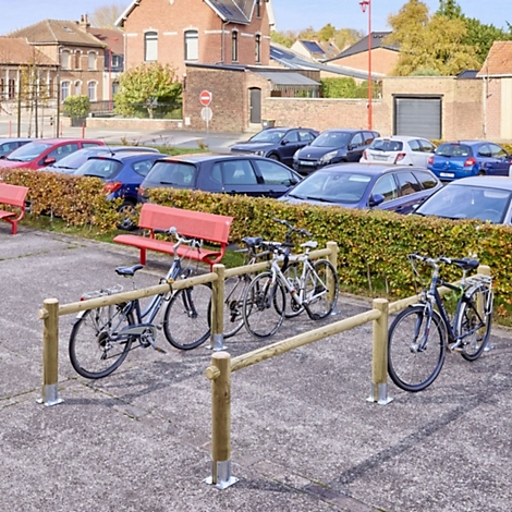 Mehrere Fahrräder stehen vor einem hölzernen Fahrradständer auf einem Parkplatz vor einer Hecke und Autos.