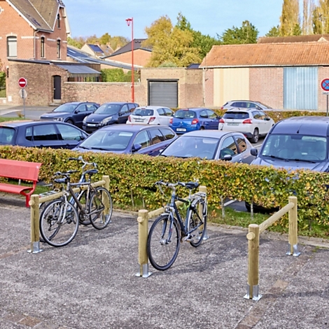 Zwei Fahrräder stehen in einem hölzernen Fahrradständer auf einem gepflasterten Platz vor einer Hecke und einem Parkplatz.