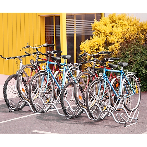 Verschillende fietsen geparkeerd in een buitenfietsenrek. Op de achtergrond een geel gebouw en groene gebieden.