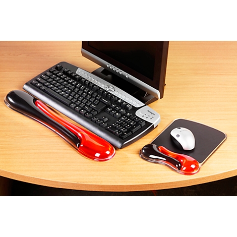 A black keyboard and silver monitor sit on a wooden desk. A red and a black wrist rest lie in front of them.