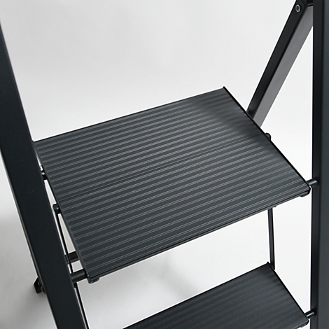 Detailaufnahme einer schwarzen Trittleiter. Die Stufen sind geriffelt, der Hintergrund ist hell.