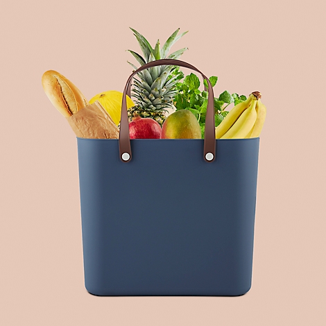 Dark blue tote bag with brown handles, filled with fruits and bread. Pineapple, bananas, mango and baguette are visible.
