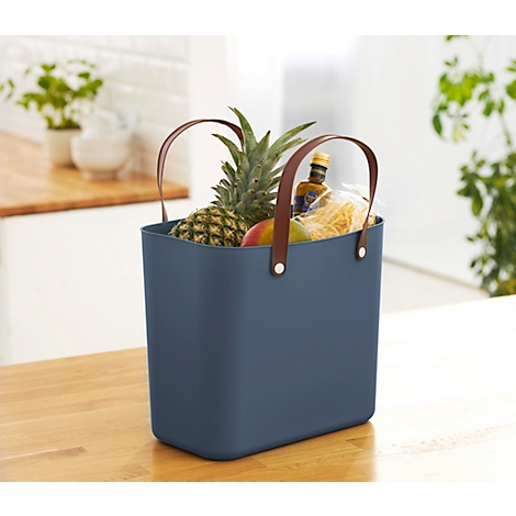 Dark blue shopping bag with brown leather handles, filled with pineapple, mango, oil and pasta. It stands on a wooden surface.