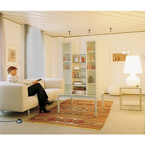 Une femme est assise sur un canapé blanc et regarde la télévision. La pièce est lumineuse avec un tapis beige et un grand abat-jour blanc à côté d'une table en verre.