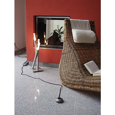 Une pièce confortable avec un sol en pierre grise. Un fauteuil en osier avec un coussin blanc est placé à côté d'une cheminée. Un livre ouvert repose sur le fauteuil. Une lampe moderne éclaire la scène.