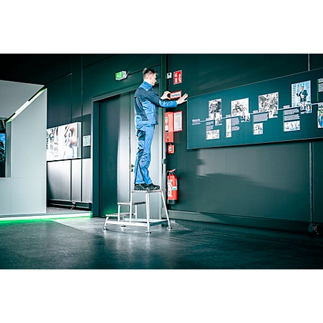 Mann montiert an einer Wand im Raum etwas, er steht auf einer kleinen Treppe. An der Wand befinden sich Bilder und Informationen.