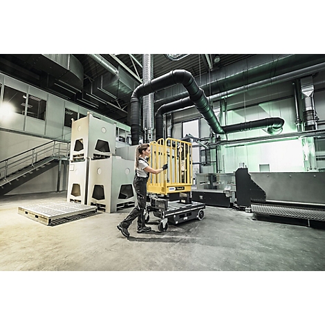 Une femme tire un chariot jaune dans un hall de production. Machines grises et tuyaux en arrière-plan.